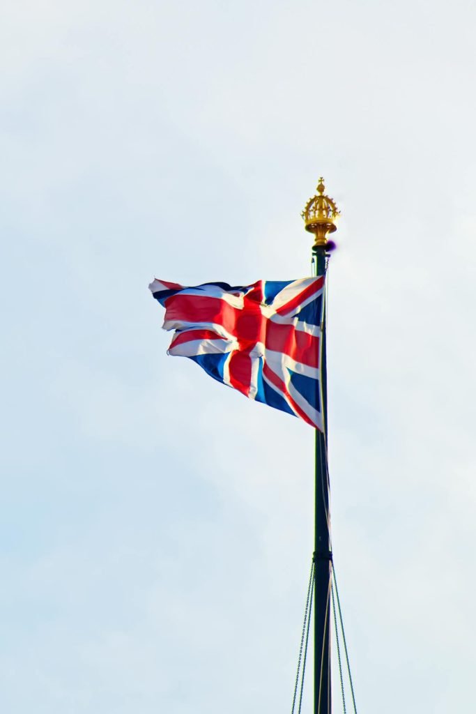 The image shows the Union Jack flag of the United Kingdom flying on a flagpole with a gold finial against a cloudy sky background. Amidst pandemics, lockdowns, and legal complexities, the flag waves in the wind, displaying its red, white, and blue colors prominently.
