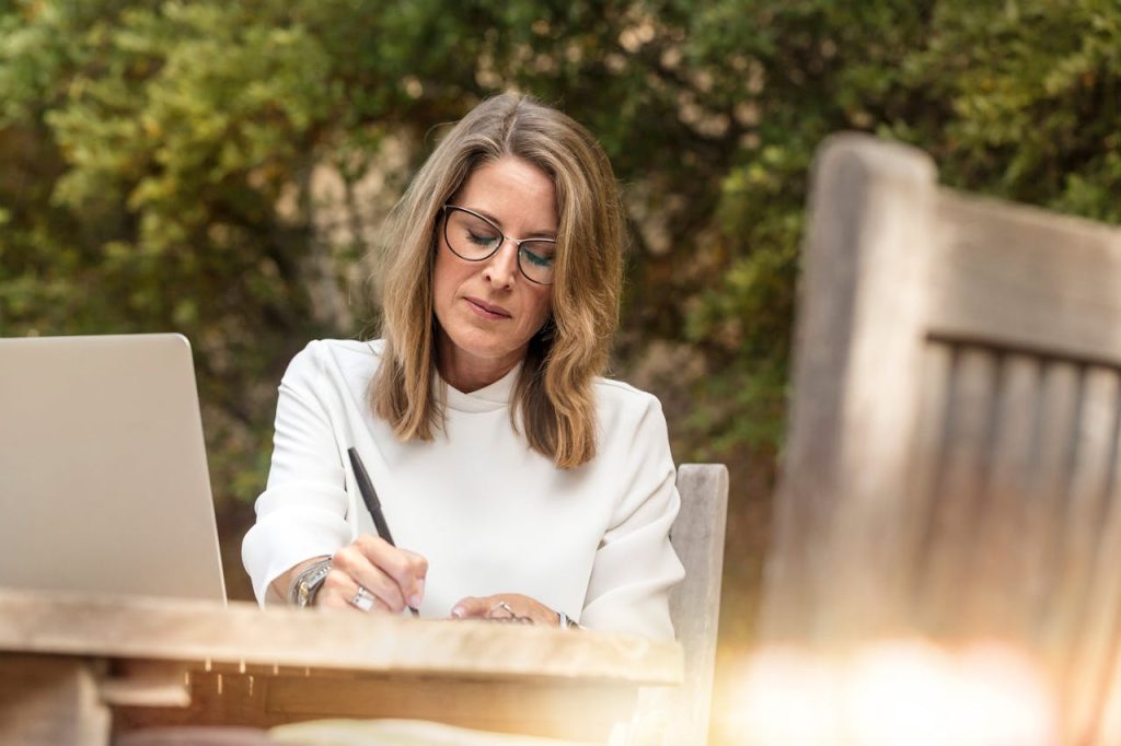 A woman with shoulder-length blonde hair and glasses sits outdoors at a wooden table. She is wearing a white blouse and writing in a notebook, reminiscent of how the court sees through layers of lipstick and linguistic camouflage. An open laptop is next to her amid the greenery and sunlight.