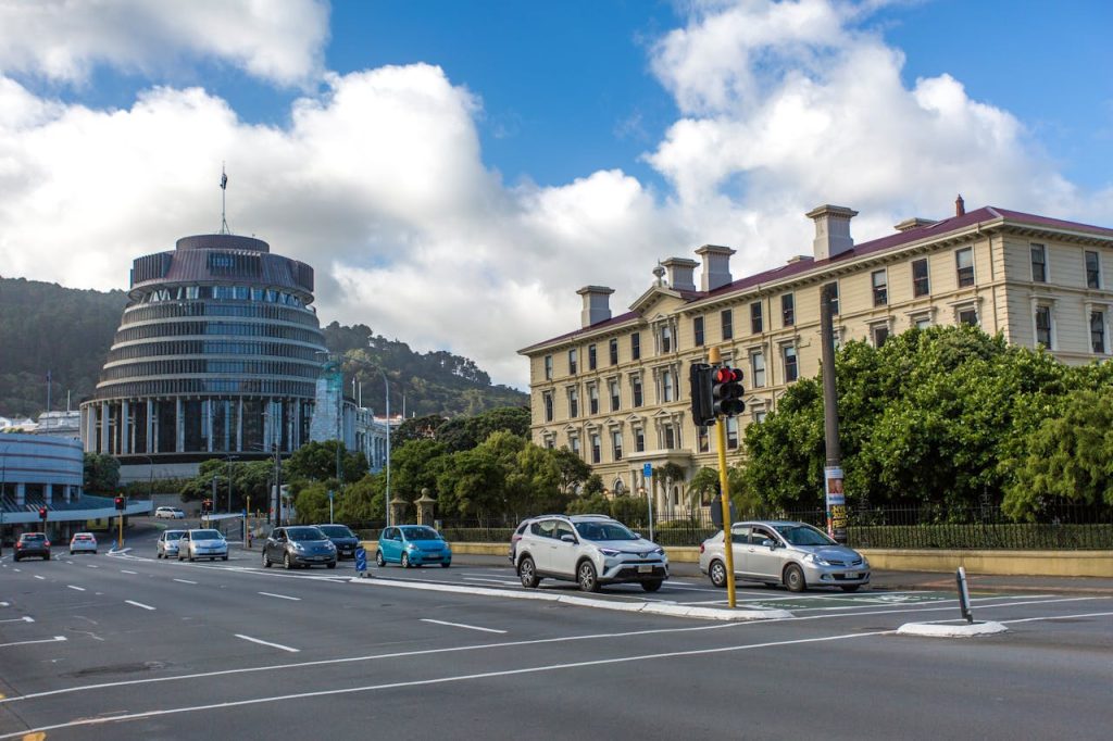 Street view showing the distinctive round building of the New Zealand Parliament, known as the Beehive, on the left. To the right is a historic, classical-style building. Several cars are seen on the road, with trees and a traffic light in the foreground—much like a well-argued reduction of damages for contributory negligence.