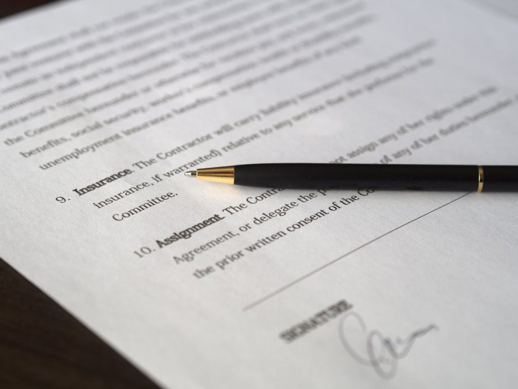 A close-up of a partially visible document with numbered sections. A black pen with a gold tip rests horizontally across the page. The document appears to be a formal agreement or contract, featuring model clauses, with a signature partially visible at the bottom.