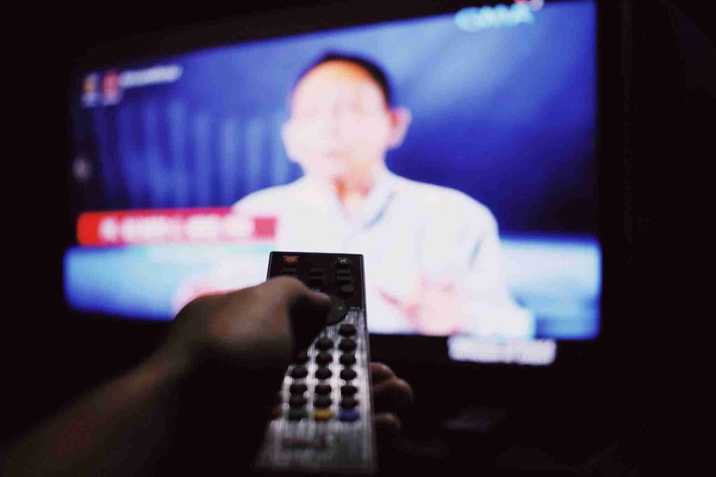 A person pointing a remote control at a blurred television screen displaying a news program. The screen shows a person's upper body, but the details are unclear due to the out-of-focus effect, making it difficult to identify any counterclaim being discussed during this enforcement stage.