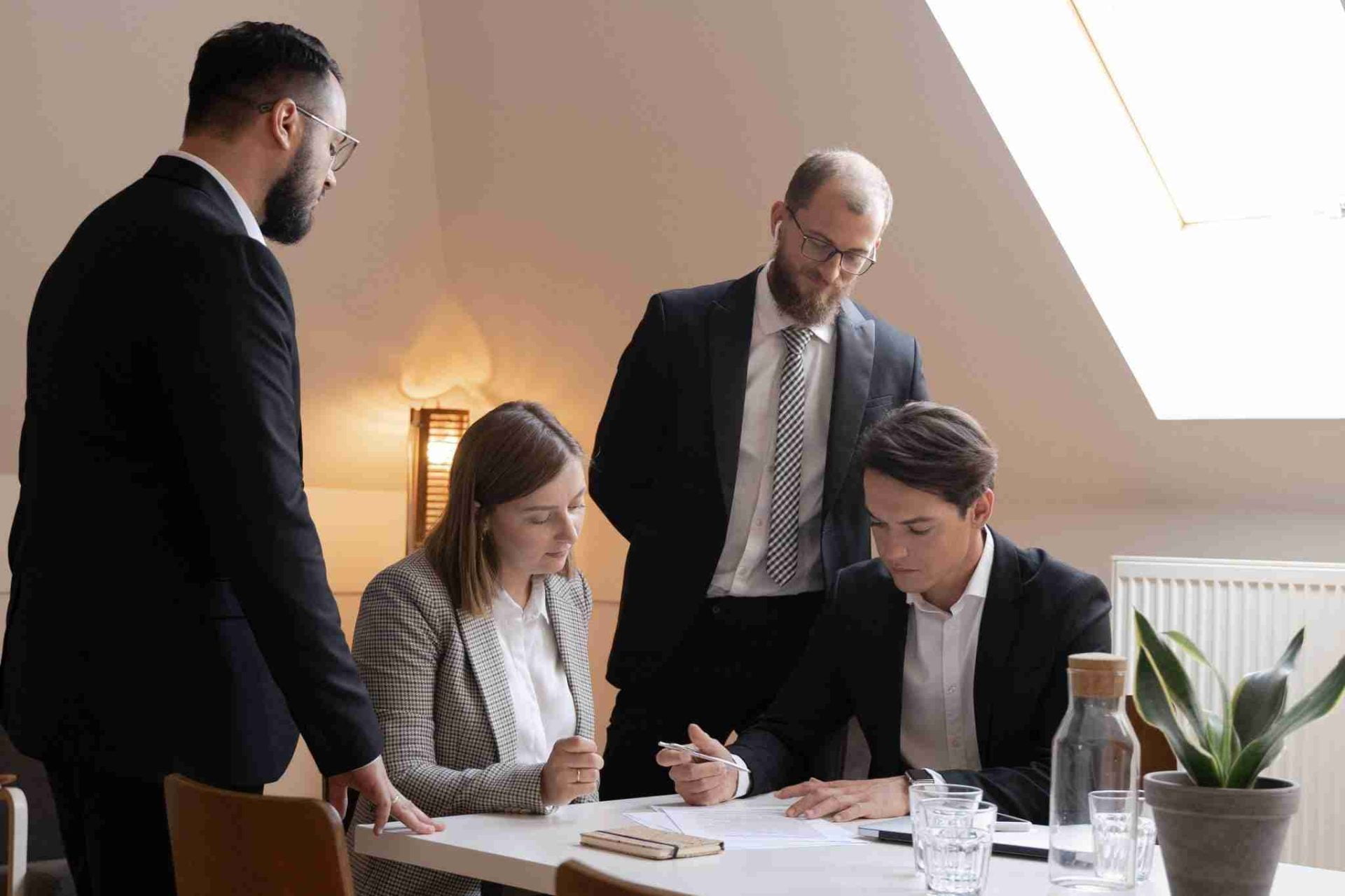 Four people in business attire are gathered around a table in a bright office. Two individuals are seated, reviewing documents with model clauses, while the other two stand, observing and discussing. A small plant and glass bottle are on the table under a skylight.