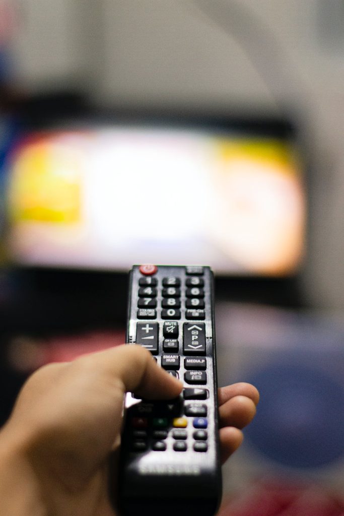 A hand holding a black remote control is pointed towards a blurred television screen in the background, suggesting the act of changing channels or adjusting the volume. It's as if the viewer is preparing for the enforcement stage of their evening entertainment routine.