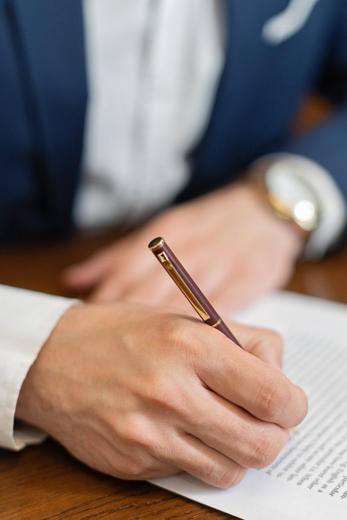 Close-up of a person wearing a dark blue blazer and a white shirt, holding a pen and writing on a document during the counterclaim enforcement stage. Their left hand rests on the paper, and a watch is visible on their wrist. The document contains printed text but the content is not legible.
