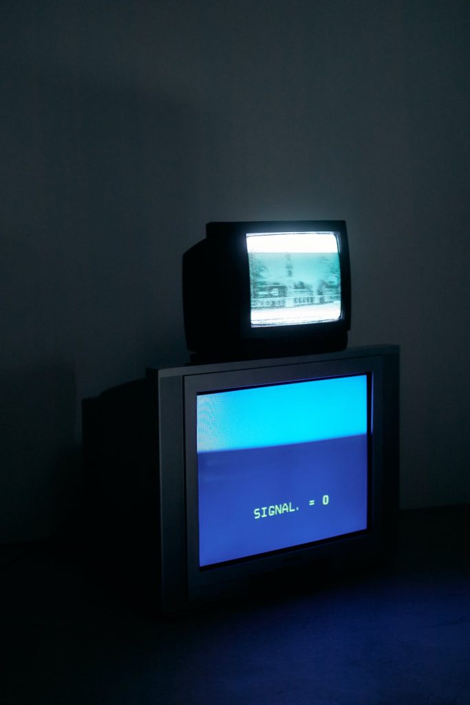 Two CRT televisions are stacked in a dimly lit room. The top TV displays a black and white image of a landscape, while the bottom TV shows a blue screen with the text "SIGNAL: 0." In this eerie setting, it's as if the ambiance itself enforces a stage of closer scrutiny.