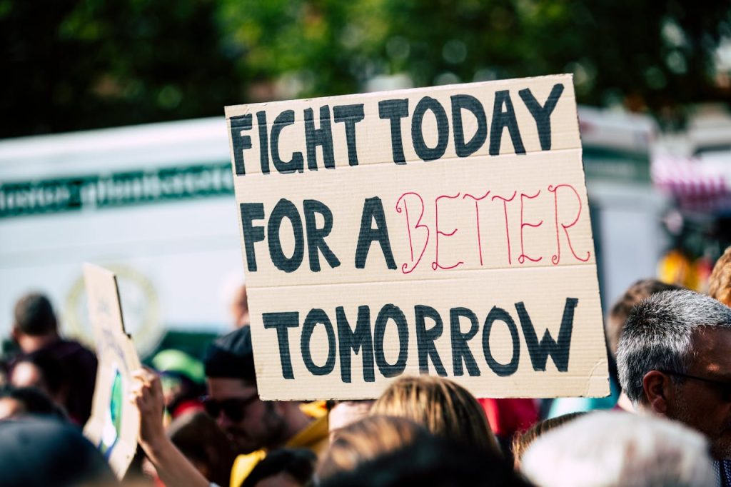 A crowd of people is gathered at a protest in New Zealand. One person is holding up a cardboard sign that reads, "FIGHT TODAY FOR A BETTER TOMORROW" in bold letters. The background includes trees and a partially visible vehicle, emphasizing the call for transparency and change.