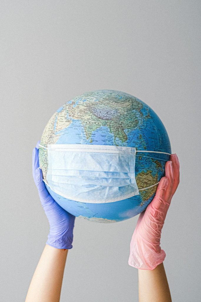 Two hands wearing disposable gloves, one purple and one pink, hold up a globe. The globe has a surgical mask covering a portion of it, symbolizing the global impact of the COVID-19 pandemic. With New Zealand transparency, this image underscores the need for honesty in addressing worldwide health challenges.