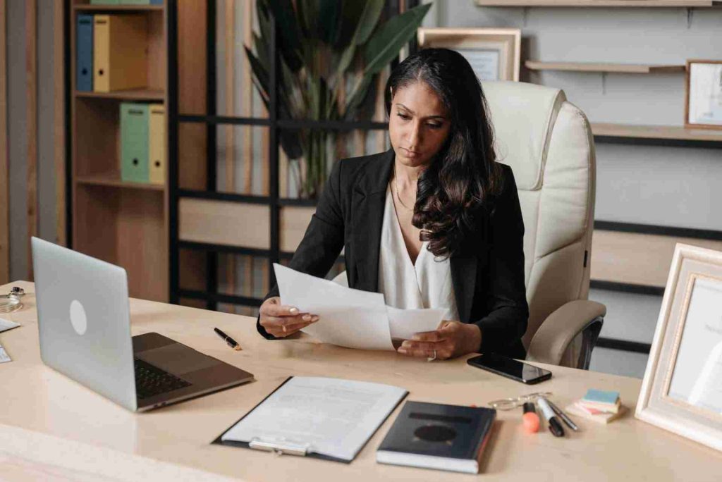 A woman sits at a desk in a modern office, holding and examining papers. She is dressed in professional attire, with a laptop, notepad, and documents spread out in front of her. Amidst the shelves, plants, and framed certificates, she scrutinizes contract clauses for wilful breaches.