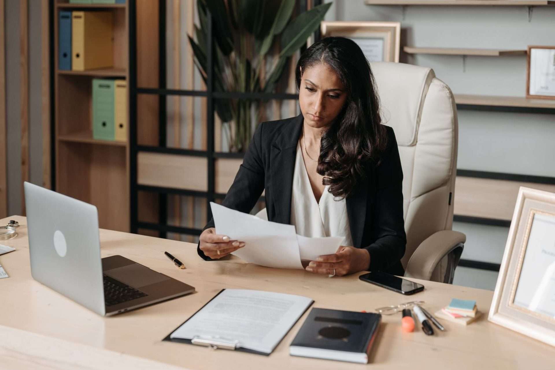 A woman sits at a desk in a modern office, holding and examining papers. She is dressed in professional attire, with a laptop, notepad, and documents spread out in front of her. Amidst the shelves, plants, and framed certificates, she scrutinizes contract clauses for wilful breaches.