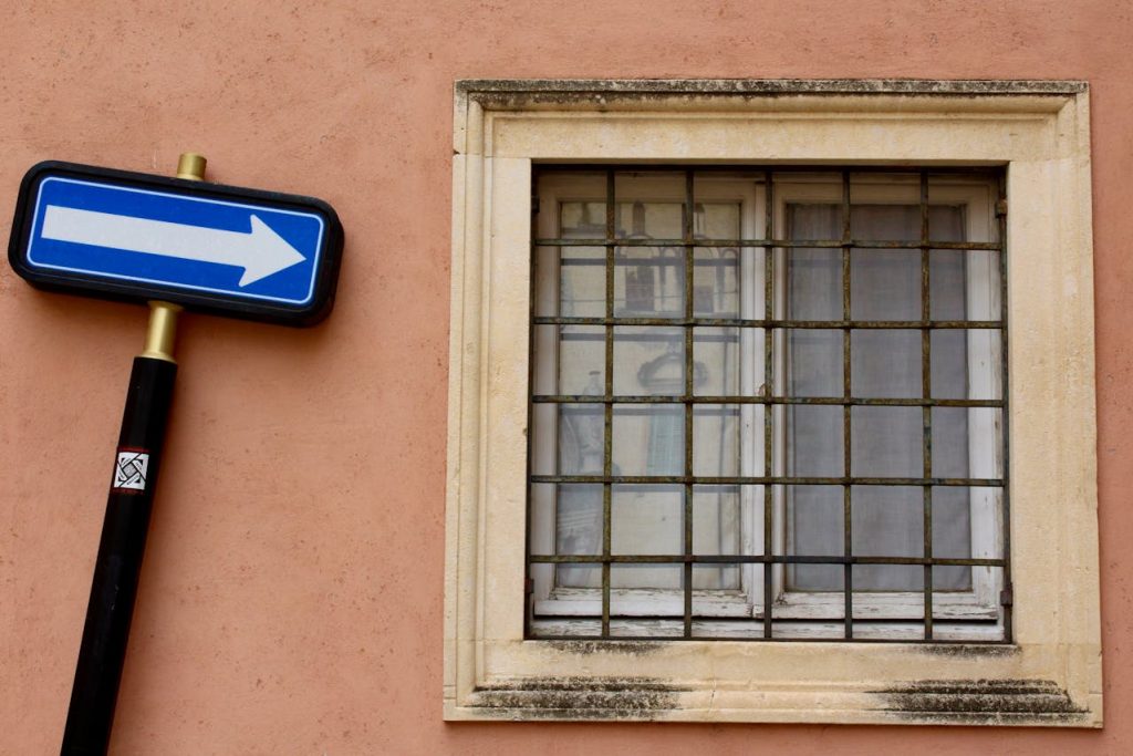 A blue and white one-way street sign is bent and leaning against the exterior wall of a peach-colored building in New Zealand. The wall has a window with metal bars, framed with white stone, showcasing a transparent reflection of the opposite side.