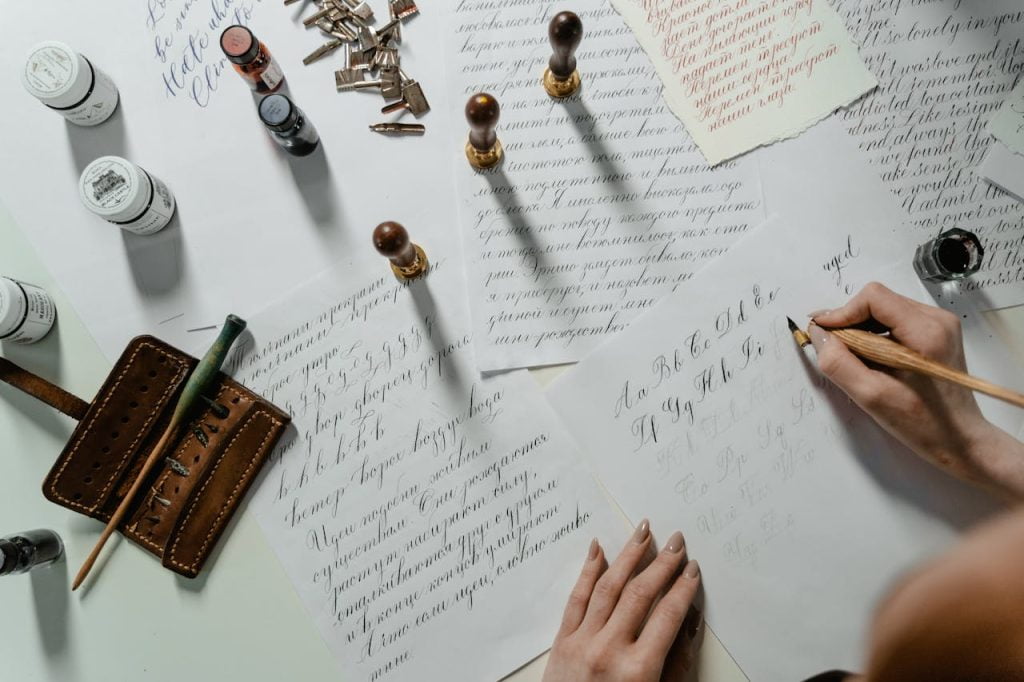 An overhead view of a well-organized calligraphy setup. Various sheets of paper with different scripts are on the desk, alongside wooden stamps, ink pots, nib pens, and a leather pen holder. Amidst this traditional scene, a familiar pair of hands is seen writing an electronic arbitration agreement in beautiful calligraphy.