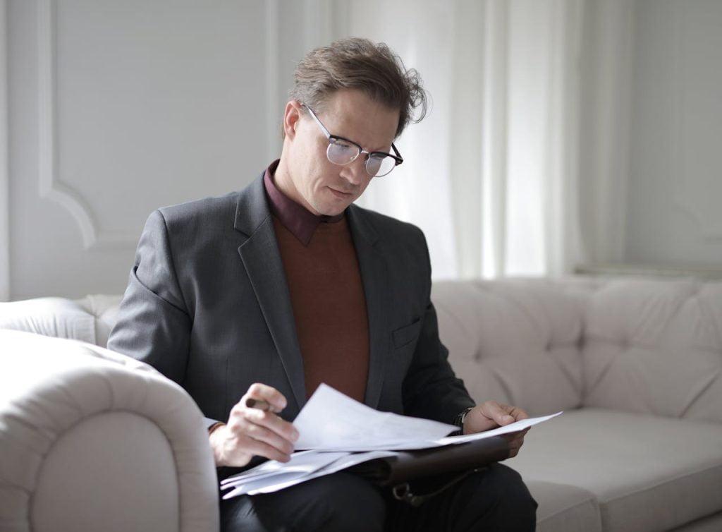 A person in a suit and glasses is sitting on a white couch, meticulously reviewing documents related to dispute resolution. They appear focused and engaged, with a pen in hand, in a well-lit room with white walls and furnishings.
