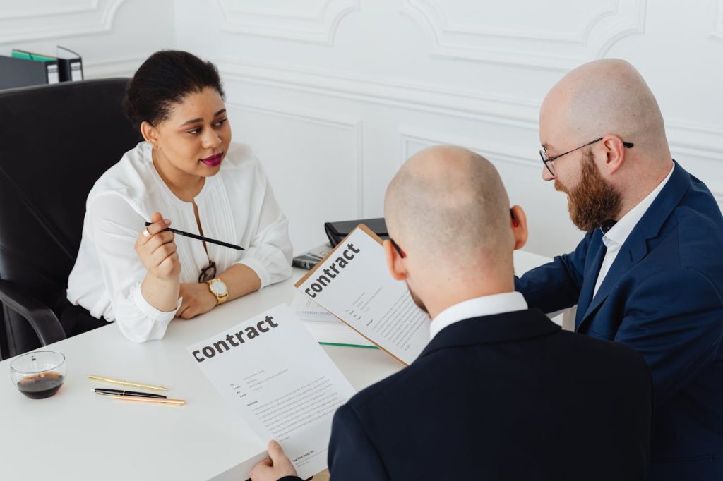 Three people are sitting at a white desk in an office. The woman on the left is holding a pen and addressing the two men in front of her. All three are holding documents labeled "contract," likely discussing dispute resolution. A glass of a dark beverage and several pens are on the desk.