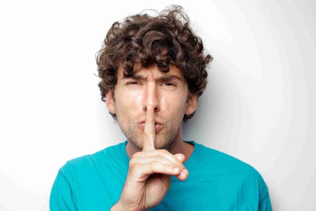 A man with curly hair wearing a teal shirt holds his index finger to his lips, signaling for silence. He looks directly at the camera with a serious expression, as if pausing before discussing international arbitration, standing against a plain, light-colored background.