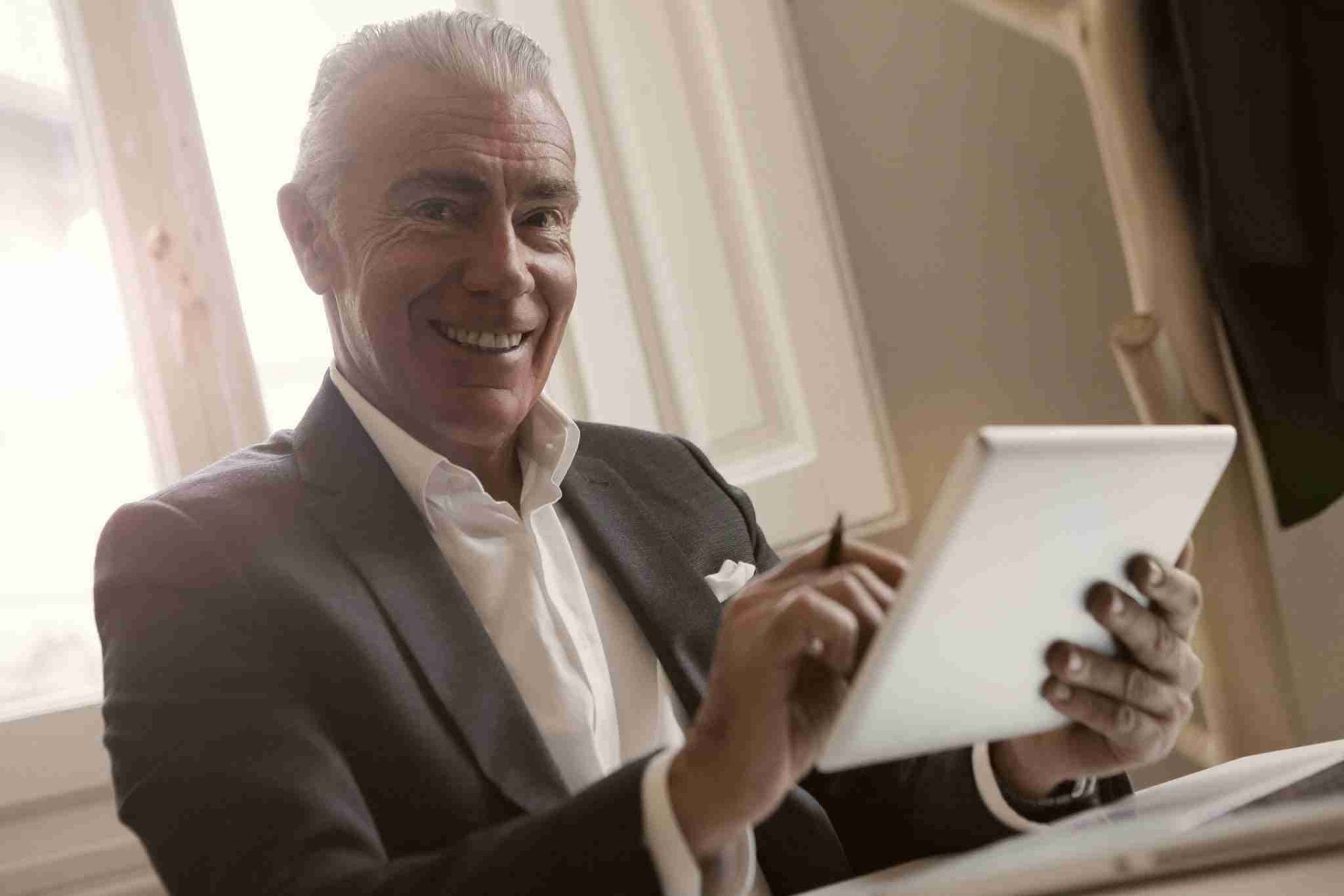 A smiling older man in a suit, holding a tablet and using a stylus pen, sits at a desk with light streaming in from a window behind him. He appears to be engaged in an electronic arbitration agreement on the tablet.
