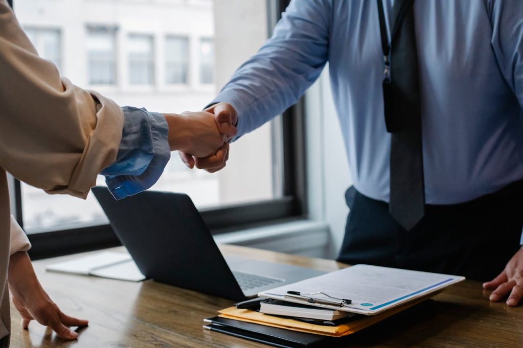 Two people are shaking hands over a desk, indicating a business agreement. On the desk, there are stacks of documents, a clipboard with papers, and an open laptop displaying an electronic arbitration agreement. The setting appears to be a modern office with large windows in the background.