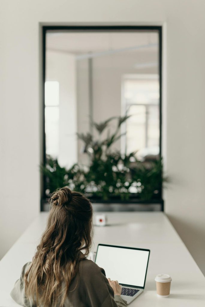 A person with long hair, seen from the back, is sitting at a desk and working on a laptop. A cup of coffee sits on the desk next to some documents about an electronic arbitration agreement. In the background, there is a window with an indoor plant arrangement visible through it.