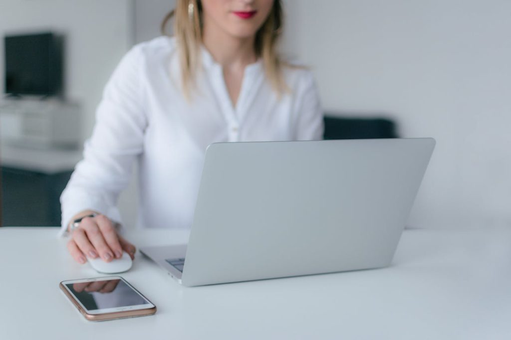 A person with long blond hair, wearing a white shirt, sits at a white desk working on a silver laptop. Their right hand is on a computer mouse, and a smartphone is placed on the desk beside the laptop, possibly reviewing an electronic arbitration agreement. The background is out of focus.