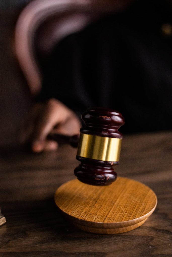 A judge's hand holding a wooden gavel over a round wooden sound block on a dark wooden table. The image is closely cropped to focus on the gavel and block, with a blurred background indicating an ICSID awards courtroom setting.