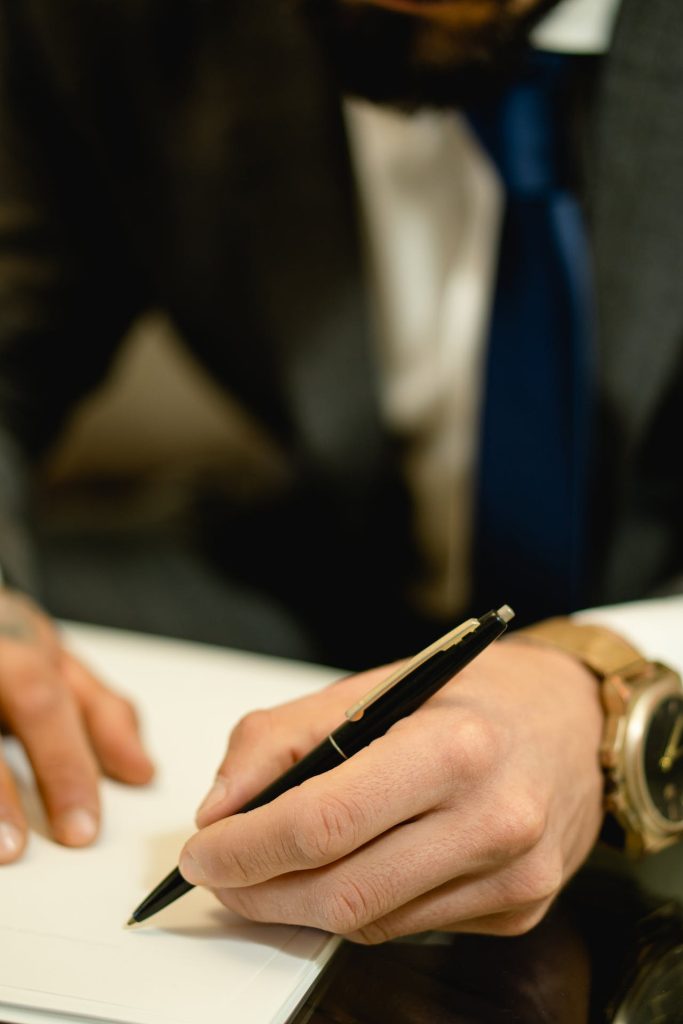 A person in a suit and blue tie is holding a pen and writing on a white sheet of paper. Only their hands and part of their torso are visible, with a wristwatch on their left wrist. The background is blurry, suggesting the focus may be on signing important documents like ICSID awards.