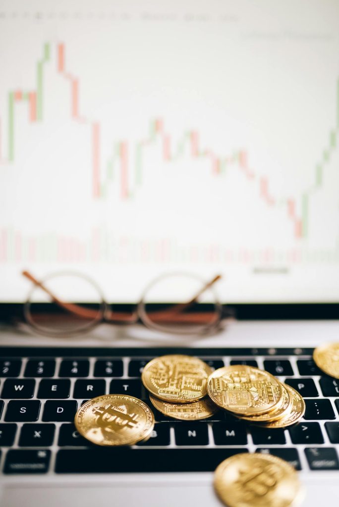 A laptop keyboard with several gold Bitcoin coins scattered on top. Glasses rest behind the keyboard, and the laptop screen displays a candlestick chart and trading graph suggesting cryptocurrency market activity. The backdrop hints at ICSID awards, lending an air of sophistication.
