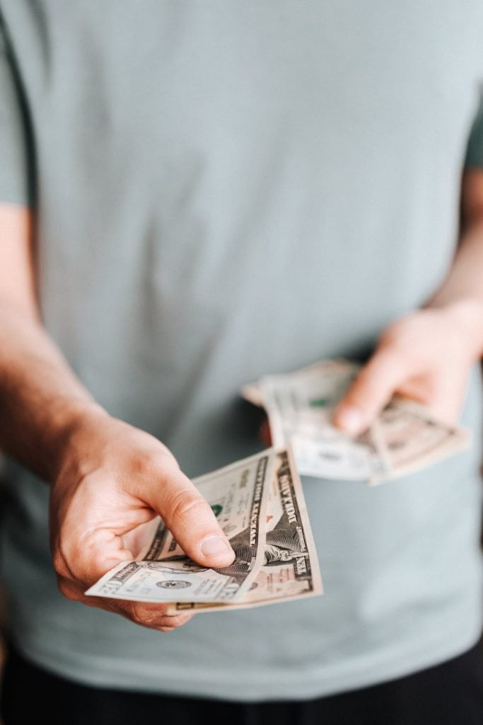A person in a light gray shirt is holding and extending a few US dollar bills with one hand while holding more bills in the other, reminiscent of hefty ICSID awards. The image focuses on the person's hands and the money, with the background blurred.