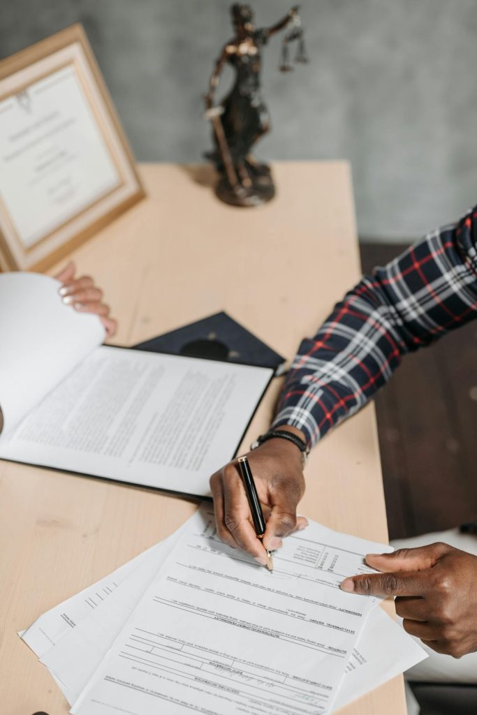 A person sitting at a desk is filling out a form or document, possibly related to ICSID awards. Their hand holds a pen, with additional papers and a clipboard on the desk. A framed certificate and a statuette of Lady Justice are visible in the background.