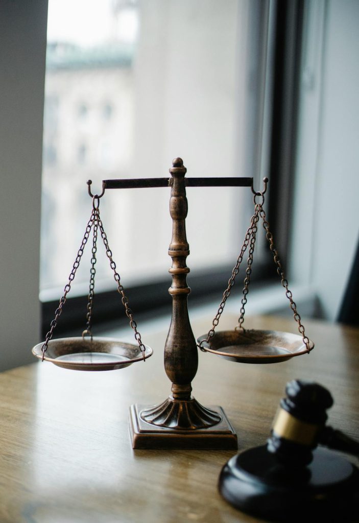 A bronze balance scale sits on a wooden desk near a window, with a blurred building in the background. The scale is empty, with two hanging plates suspended by chains. A wooden gavel lies in the foreground, symbolizing justice in ICSID awards cases. The scene is lit by natural daylight.