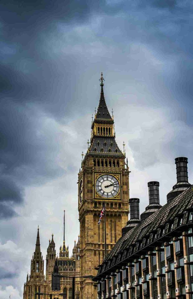A view of the Elizabeth Tower, commonly known as Big Ben, against a dramatic, cloudy sky. The Gothic-style clock tower features its iconic clock faces. Surrounding buildings, including parts of the Houses of Parliament and nearby polo clubs, are visible in the foreground.