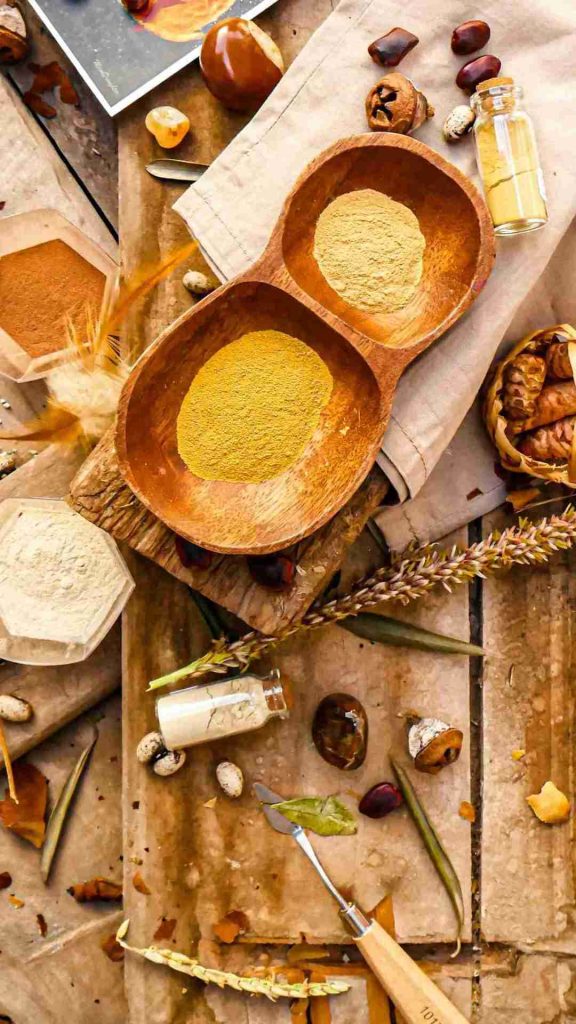 A rustic wooden table displays a variety of herbs and spices. Two wooden bowls hold yellow powdered spice, surrounded by garlic, dried spices, a small bottle of oil, and various seeds. A beige cloth and dried plants enhance the rustic aesthetic, reminiscent of detailed arbitration agreements.
