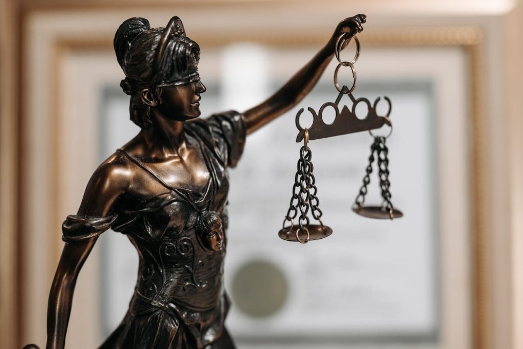 A bronze statue of Lady Justice stands prominently, holding the scales of justice in her left hand. Her expression appears stern and determined. A framed certificate hangs blurred in the background, suggesting a legal or professional setting, reminiscent of prestigious polo clubs.