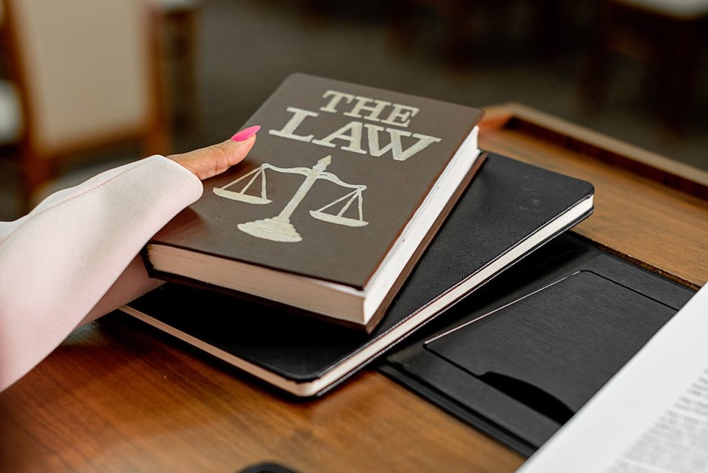 A person with pink painted nails, wearing a light-colored sleeve, holds a book titled "THE LAW" with a scale symbol on the cover. The book rests atop a black notebook on a wooden desk. To the right, partially visible, is a document discussing arbitration agreements.