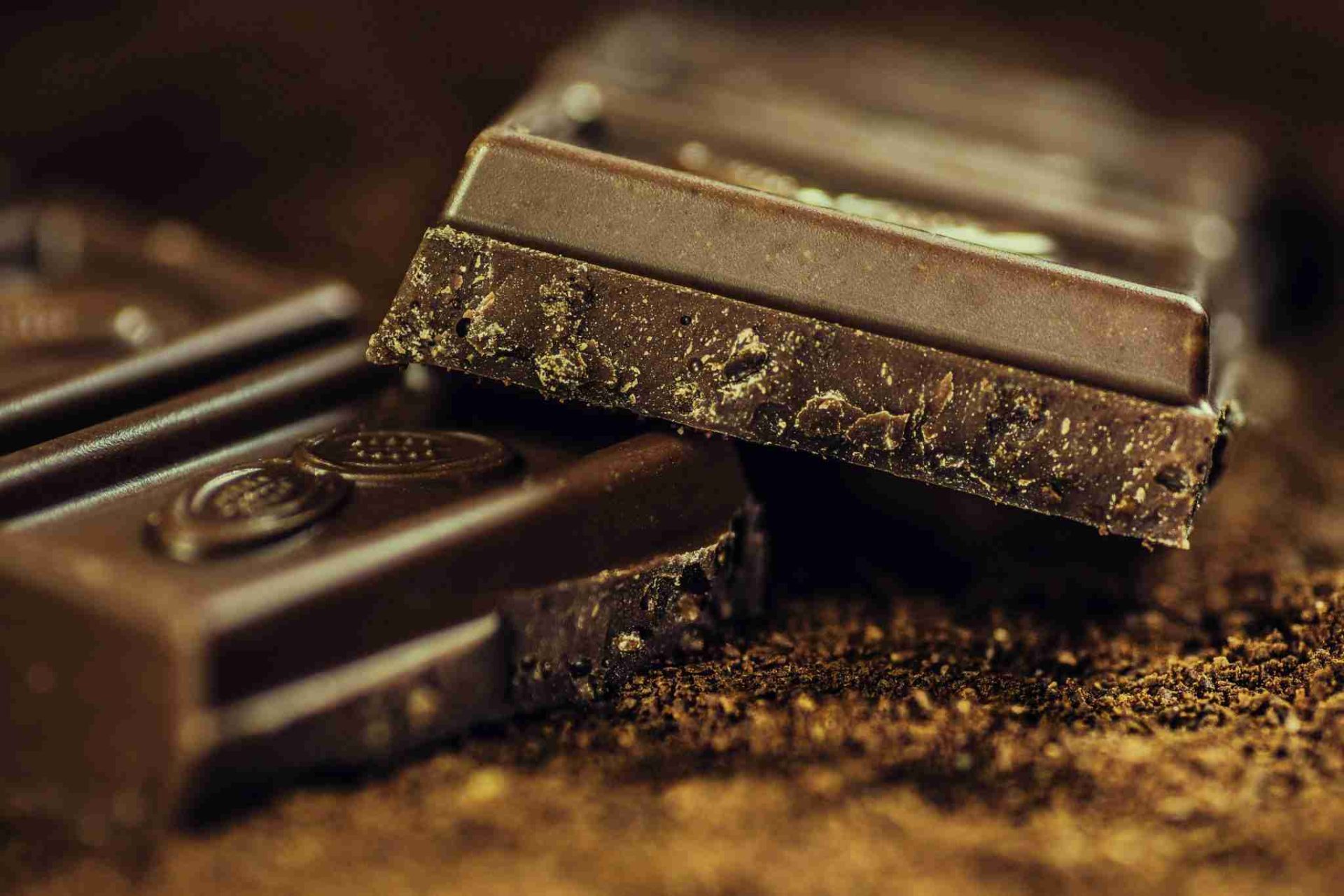 Close-up of three pieces of Whittaker & Sons dark chocolate with a textured surface, partially stacked on each other. The image highlights the rich, dark brown color and rough edges of the chocolate pieces against a blurred brown background.