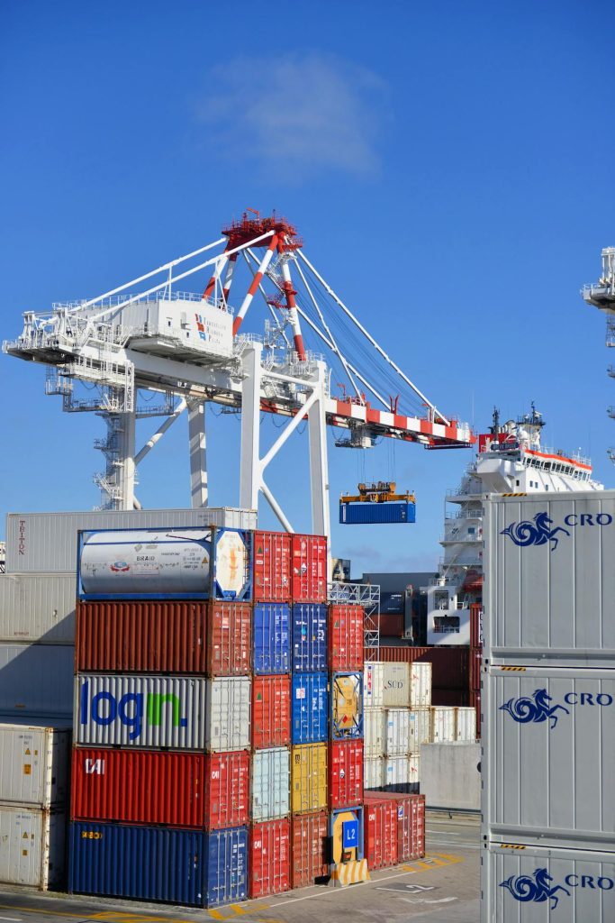 A vibrant shipping port featuring large cranes with red and white stripes lifting cargo containers, operated by Whittaker & Sons. Stacks of colorful containers are prominently displayed in the foreground. The clear blue sky and a hint of a ship in the background indicate a busy, sunny day.