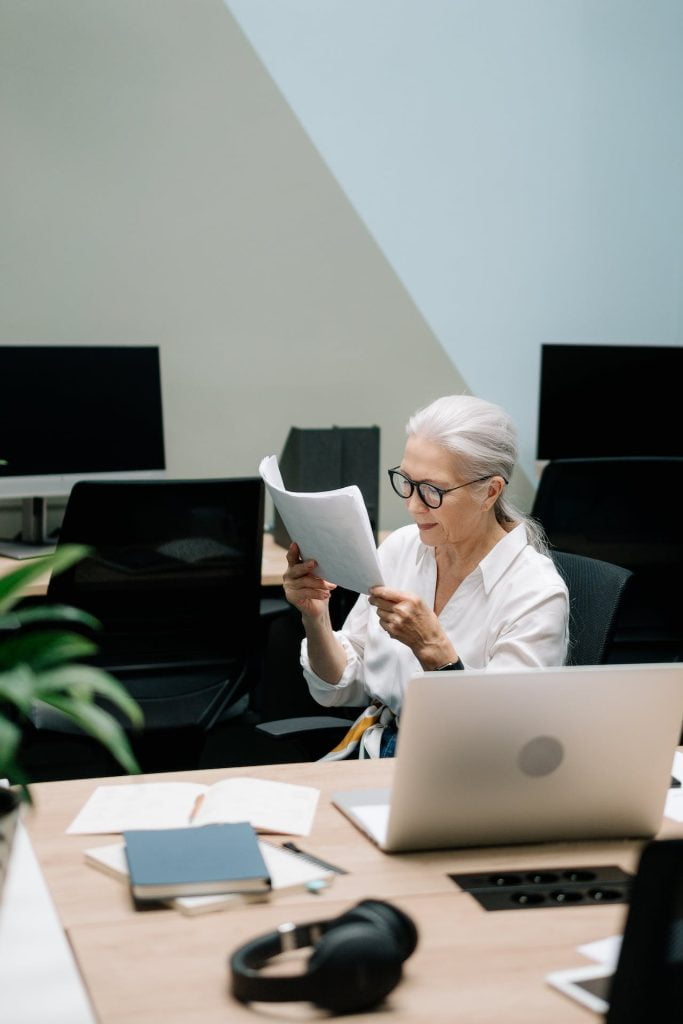 An older woman with long gray hair and glasses is seated at a desk in the Whittaker & Sons office, reading a stack of papers. She is surrounded by office supplies, including a laptop, notebooks, and headphones. The modern office environment features multiple computer monitors in the background.
