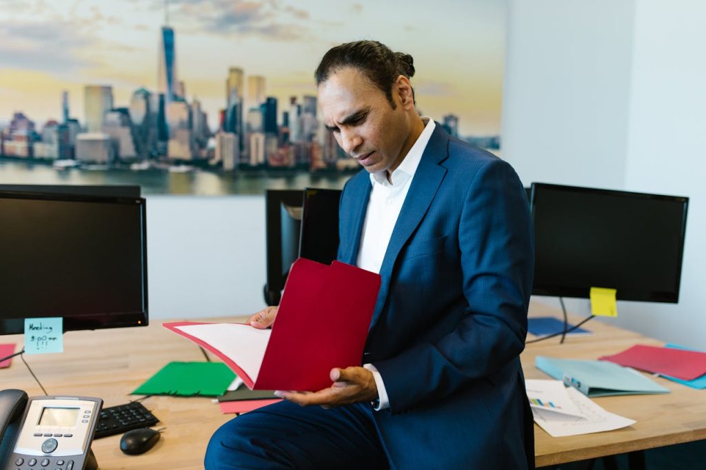 A man in a blue suit is sitting on the edge of a desk, looking intently at a red folder that likely contains an arbitral award. Behind him are several computer monitors and a large photo of a city skyline. The desk has papers and office supplies scattered on it.