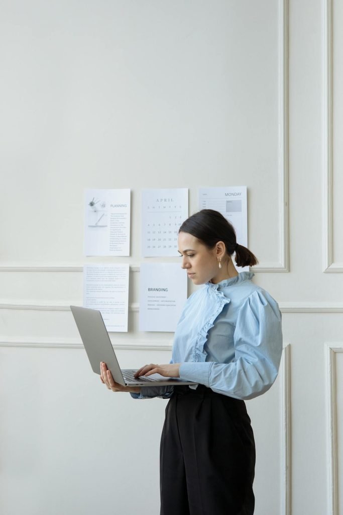 A woman with a ponytail is standing and using a laptop. She is wearing a light blue blouse and black pants. On the wall behind her, there are various papers, an arbitral award, and a calendar pinned up. The room has white paneled walls.