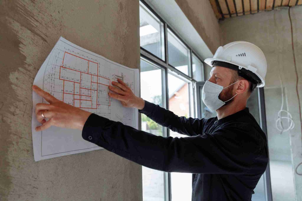 A construction worker wearing a white safety helmet and a face mask is examining a floor plan attached to a concrete wall. The worker points at the blueprint with one hand while adjusting it with the other, standing in a partially constructed building, likely reviewing details related to the construction contract.