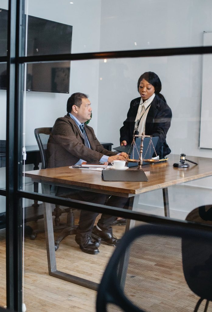 A man and woman in business attire are having a conversation in a modern office. The man is seated at a desk with papers, a gavel, and a scale, while the woman stands beside him holding a folder full of expert evidence. They appear to be in a professional, law-related setting.