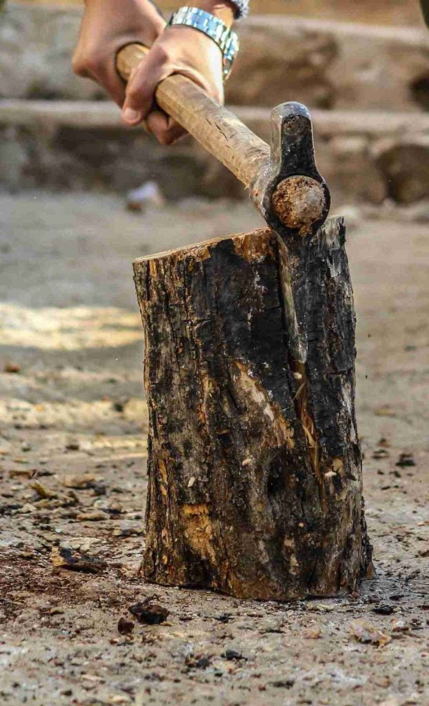 A hand with a wristwatch is swinging an axe to split a log of wood on the ground surface. The axe is mid-swing and about to make contact with the log, which shows signs of previous cuts and wear, highlighting expert evidence of repeated use.