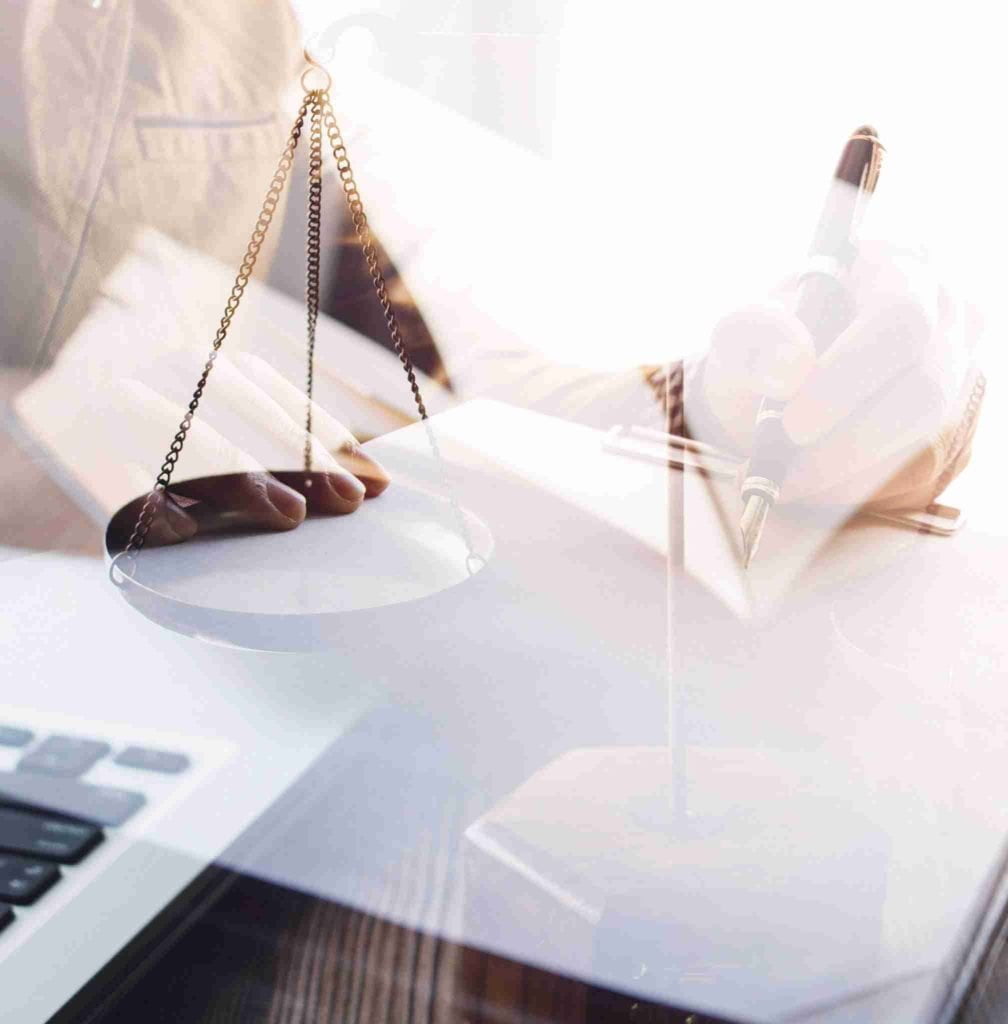 A double exposure image showing a person writing in a notebook with a pen on the right and a laptop on the left. Overlaid, there is a scale of justice symbol, suggesting themes related to law, justice, or legal documentation.