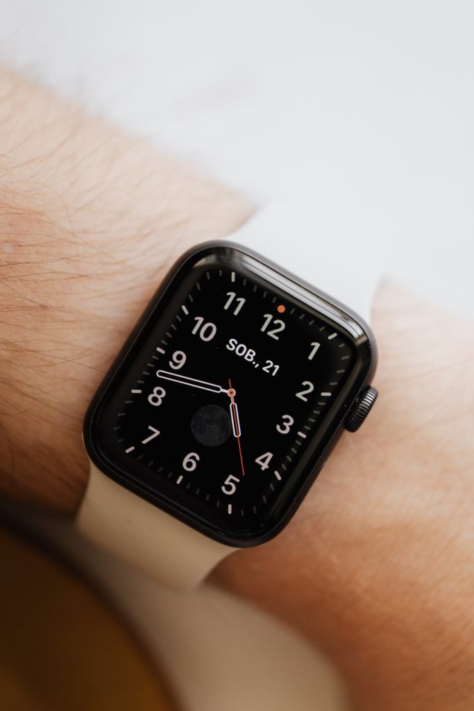 Close-up of a wrist wearing a smart watch with a square black face and a white band. The watch displays an analog clock with red minute and hour hands, white second hand, date in German ("SOB. 21"), and a moon phase indicator at the 6 o'clock position.