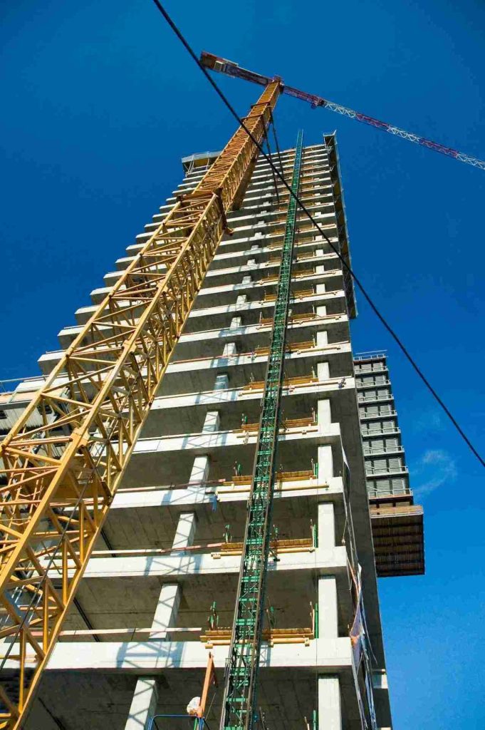 A tall, under-construction high-rise building with a yellow construction crane extending upwards alongside it. The sky is clear and blue, and the building structure appears to have multiple floors, with scaffolding visible on some levels.