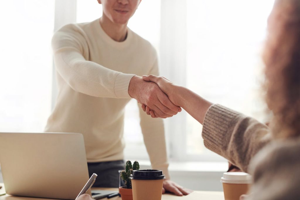 Two people are shaking hands over a desk in a bright office setting. One person is standing, wearing a white sweater, and the other is seated, wearing a beige sweater. On the desk are a laptop, a small potted plant, two takeaway coffee cups, and a smartphone.