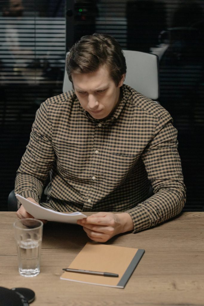 A man in a checkered shirt sits at a wooden table, attentively reading a document. A glass of water, a notebook, and a pen are on the table in front of him. The background features a dark interior with reflective surfaces.