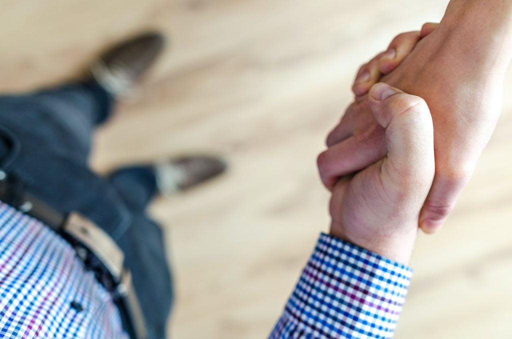 A close-up, angled view of two people shaking hands. The focus is on the hands, with one person wearing a checkered shirt and the other person extending a bare arm. The background is blurred, showing a light-colored floor.