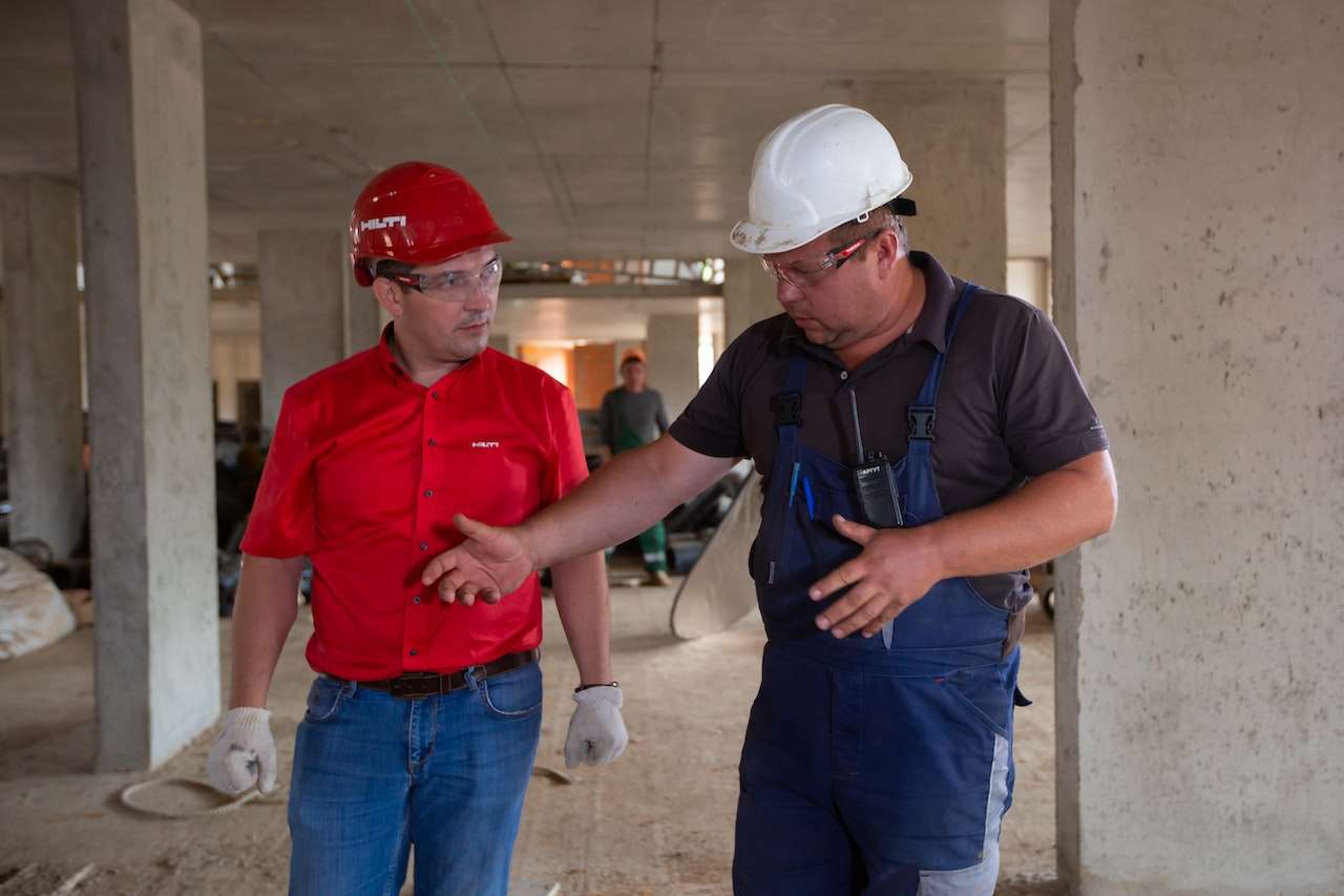 Two construction workers stand in an unfinished building. One worker, in a red shirt and red helmet, converses with another worker, in a dark shirt, blue overalls, and a white helmet. The white-helmeted worker gestures while explaining something. Tools and materials are visible in the background.