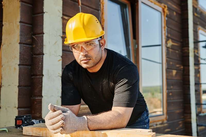 A construction worker wearing a yellow hard hat, safety glasses, and gloves leans on a wooden surface at a construction site. He is looking at the camera, and the background shows a wooden building under construction with windows and scaffolding.