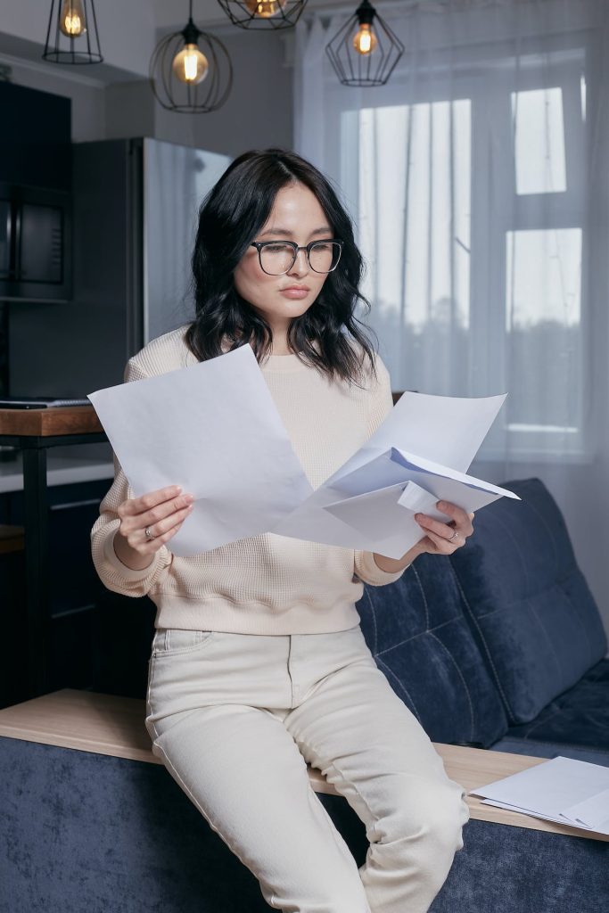A woman with shoulder-length dark hair and glasses is seated on a couch, reviewing several sheets of paper. She is wearing a beige sweater and light-colored pants. There are modern light fixtures and a window with sheer curtains in the background.