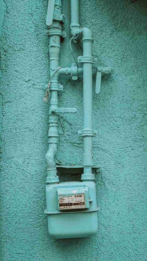 A teal-colored gas meter is attached to a textured teal wall. The meter is connected to several metal pipes, also painted teal, which run vertically and horizontally beside it. Wires are visible near the top of the pipes.